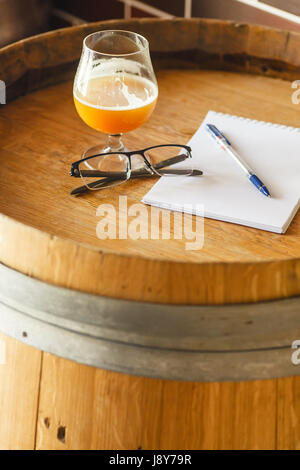 Bicchiere di birra di frumento con un paio di occhiali, una penna e un blocco note in piedi su un barile di legno in una fabbrica di birra Foto Stock