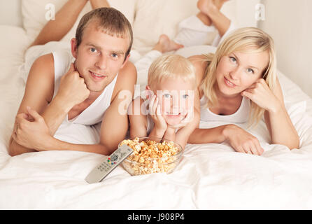 La famiglia felice: padre, madre e figlio rilassante, guardare la TV e mangiare popcorn nel letto di casa Foto Stock