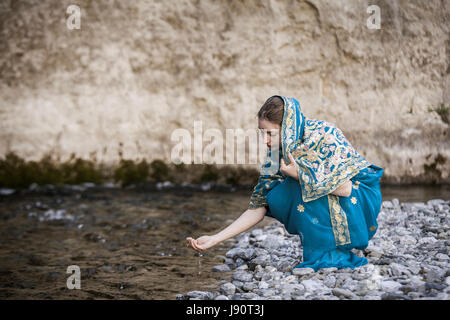 La ragazza in sari indiani si siede al piccolo fiume e bevande dalle mani Foto Stock