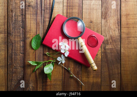 Foto del notebook rosso, Magnifier, Guarnizione cera, herry fiori e foglie verdi su vintage sfondo di legno. Elementi di cancelleria. Foto Stock