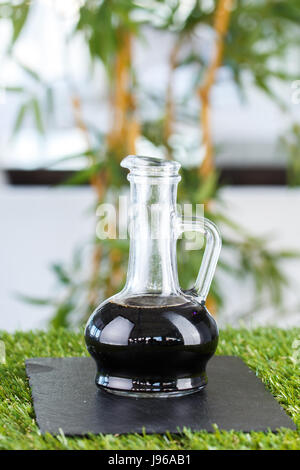 Salsa di soia scura in un decanter in vetro su uno sfondo di bambù. La salsa di soia sull'erba. Foto Stock