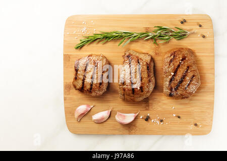 Tre fette di carne alla brace, filetti di manzo, ripresa dall'alto su una tavola di legno con un rametto di rosmarino, 4 spicchi di aglio, sale e pepe Foto Stock