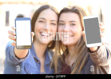 Vista frontale di due amici felici che mostra entrambi vuoti smart phone schermate in strada con una sfocatura sullo sfondo Foto Stock