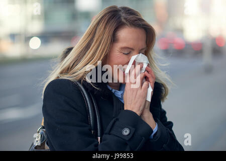 Donna con un lavoro stagionale freddo inverno soffia il naso in un fazzoletto o tessuto come lei cammina giù per una strada urbana in una salute e concetto medico Foto Stock