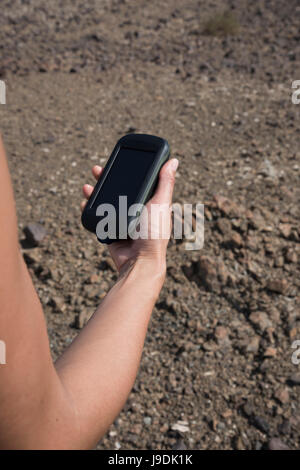 Donna che utilizza un dispositivo GPS portatile con montagne aride sullo sfondo durante un trekking, concentrandosi sull'unità GPS per la navigazione Foto Stock