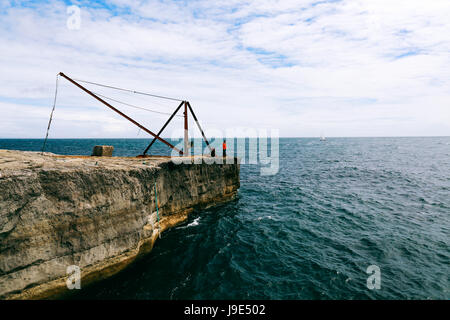 Giovane uomo si erge sulla roccia con acciaio arrugginito Fisherman's gru vicino a Portland Bill Lighthouse su Jurassic Coast Foto Stock