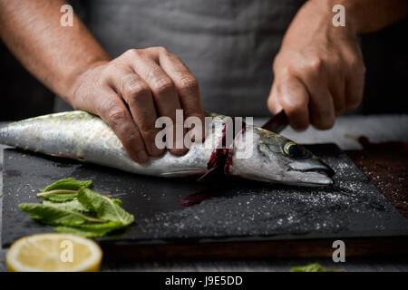 Primo piano di un giovane uomo caucasico il taglio della testa di un crudo fresco di sgombro con un coltello su un vassoio di ardesia posto su una tavola in legno rustico o top Foto Stock