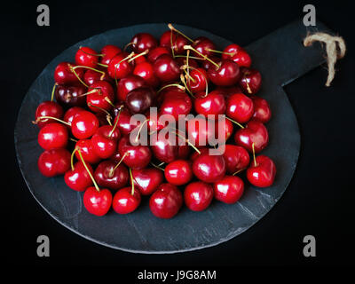 Mix di ciliegie mature preparato sul round-pietra sagomata. Luminose frutta rossa su uno sfondo nero. Foto Stock