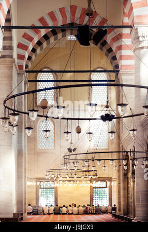 Persone sedute in preghiera all'interno di una moschea storica, che mostra archi ornati, luci sospese e architettura islamica sotto luci soffuse. Foto Stock