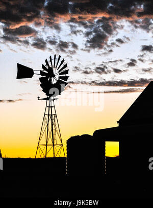 Un australiano agriturismo con mulino a vento in silhouette contro un tramonto chiazzata del cielo. Foto Stock