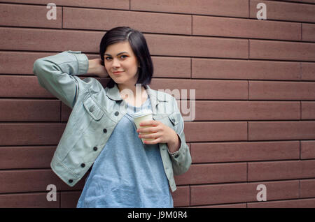 Sorridente ragazza brunnete con gallerie di orecchie in un beige giacca di pelle con una tazza di caffè in piedi di fronte a un muro di mattoni. Foto Stock