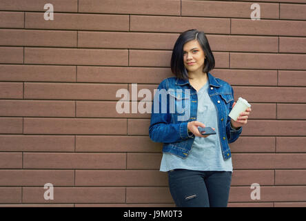 Bella bruna giovane donna con gallerie di orecchie in un jeans blu giacca con una tazza di caffè e lo smartphone in piedi di fronte a un muro di mattoni. Foto Stock