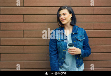 Bella bruna ragazza con gallerie nelle orecchie in un jeans blu giacca con una tazza di caffè in piedi di fronte a un muro di mattoni. Foto Stock