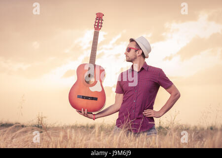 L'uomo con il cappello e occhiali da sole tenendo la chitarra acustica nel campo di grano con il tramonto e le nuvole in background. Musica, Arte, Stile di vita e il concetto di libertà Foto Stock