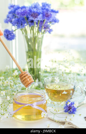 Il miele in vasetti di vetro e la tazza di tè bianco con fiori di primavera e di Fiordaliso sul davanzale. Profondità di campo. Foto Stock