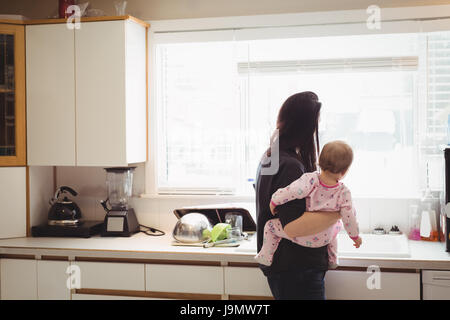 Vista posteriore della madre e del bambino guardando attraverso la finestra della cucina a casa Foto Stock