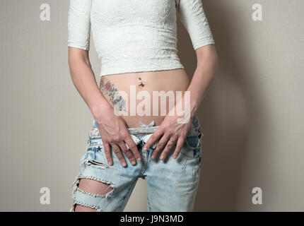 Giovani caucasici slim donna che indossa corto superiore a maglia con i pollici in linea cintura di aprire il taglio jeans blu, rivelando mutandina top, addome e tatuaggio. Regno Unito Foto Stock