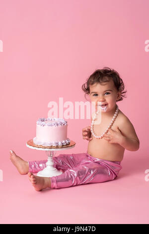Un felice, un anno di età, baby ragazza seduta con una torta. Indossa gambali rosa, un filo di perle e ha la glassa sul suo mento. Foto Stock