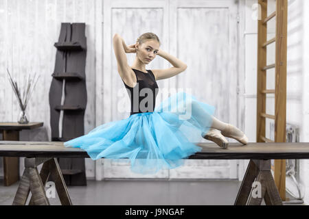 Attrective ballerina siede sul tavolo di legno sulla parete di luce sullo sfondo in studio. Indossa un nero body con un tutu ciano e pointe scarpe. Foto Stock