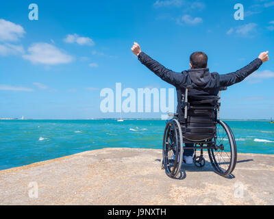 Solo disabili giovane uomo seduto in una sedia a rotelle e guarda al mare. Foto Stock