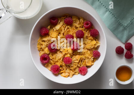 Cereali per la prima colazione di cornflakes e lamponi e miele e latte sul tavolo bianco Foto Stock