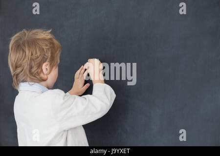 Vista posteriore del giovane ragazzo vestito come insegnante e scrive sulla lavagna nera Foto Stock