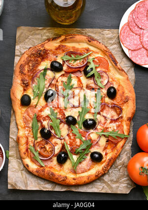 Pizza con pomodoro, salumi e olive su carta su sfondo scuro, vista dall'alto Foto Stock
