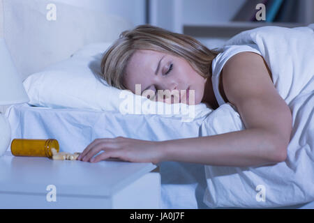 Giovane donna che dorme nel letto dalla fuoriuscita di una bottiglia di pillole sul tavolo a casa Foto Stock