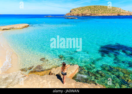 Unidentified giovane donna in costume da bagno in piedi su una roccia e posa contro la bellissima Cala Comte beach, isola di Ibiza, Spagna Foto Stock