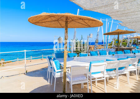 Spiaggia di Cala Nova, isola di Ibiza - 20 Maggio 2017: Bianco tavolo e sedie con cuscini blu sulla terrazza ristorante sulla spiaggia di Cala Nova con belle vie del mare Foto Stock