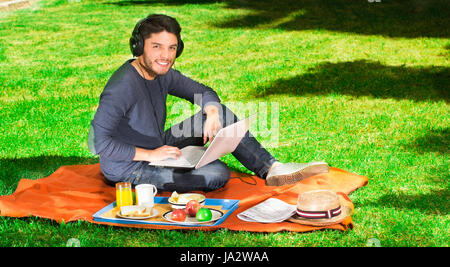 Giovane uomo felice godendo picnic nel parco mentre egli utilizza il suo computer di indossare una cuffia Foto Stock