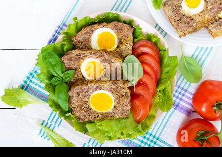 Polpettone farcito di uova sode con pomodoro fresco e foglie di lattuga su una piastra su un bianco tavolo di legno. Foto Stock