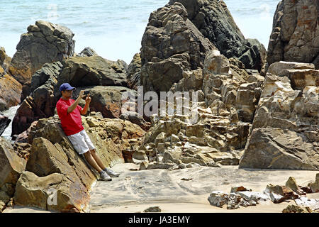 Turistica prendendo foto utilizza lo smartphone di rocky, resistente e pungenti e tipo di scultura costa al Arambol Beach in Goa, India Foto Stock
