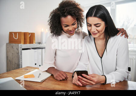 Due attraenti multirazziale giovani imprenditrici seduti ad un tavolo in ufficio la lettura di un messaggio di testo su un telefono cellulare con un sorriso Foto Stock