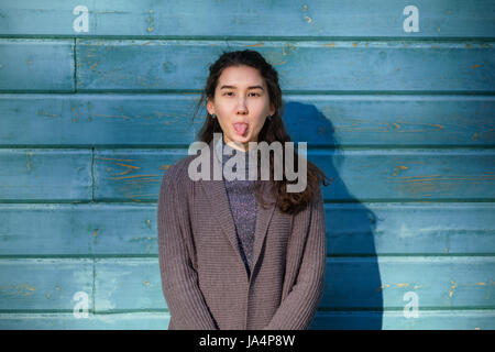 Una bella ragazza asiatica stolti intorno e mostra la sua lingua. Essa si erge dal legno parete blu. Indossa una giacca calda. Foto Stock