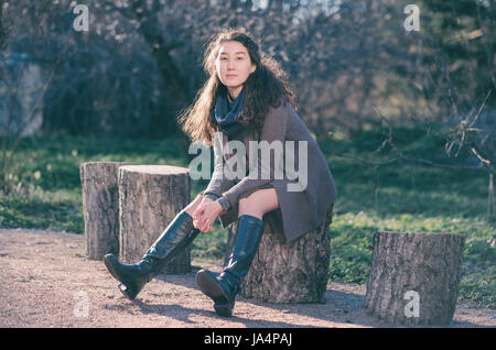 Una bella ragazza asiatica si trova in un parco su un moncone e stretching rilassa le gambe in avanti. Godetevi il silenzio nella vita della città. Foto Stock