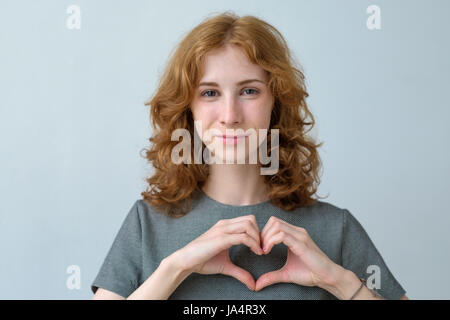 I Capelli rossi ragazza con lentiggini mostra la silhouette di cuore con l aiuto delle mani. La cura per la salute o un suggerimento di un rapporto romantico Foto Stock