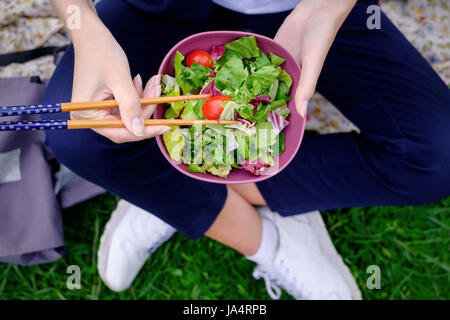 Una ragazza si siede sul prato e mangia un verde insalata fresca con bacchette per sushi. Una sana dieta su alimenti vegetali. Foto Stock