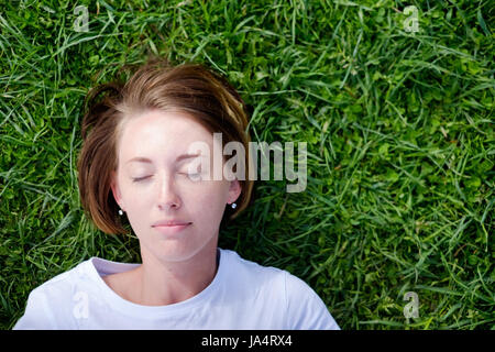 Una bellissima ragazza con le lentiggini giace sull'erba verde con gli occhi chiusi e poggia Foto Stock