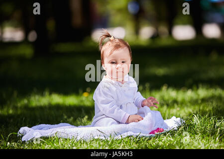 Carino baby ragazza seduta sul prato verde nel parco della città a caldo giorno d'estate. Foto Stock