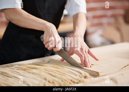 Interni orizzontali crop colpo di donna per affettare impastare e preparare gli spaghetti nel ristorante caffetteria. Fare la pasta. Fare la pasta nel ristorante. Irriconoscibile wo Foto Stock