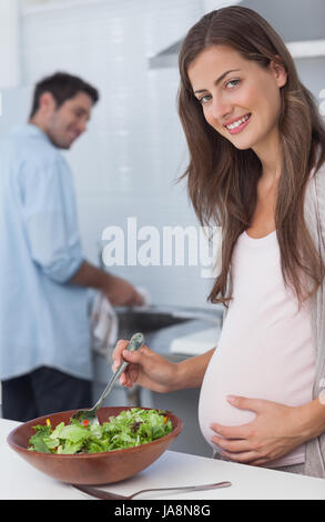 Donna incinta preparare una insalata in cucina con suo marito lavando i piatti dietro Foto Stock