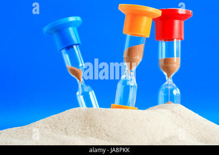 Concetto di tempo. Tre hourglasses colorati in piedi nella sabbia su sfondo blu Foto Stock