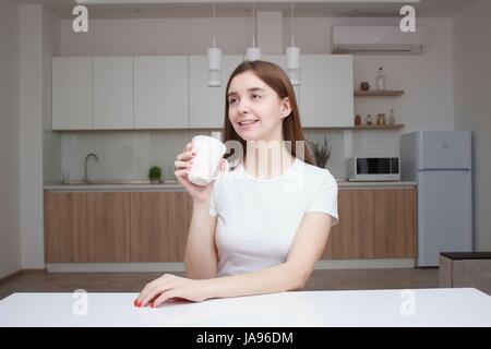 Donna sorridente seduta in cucina con la tazza di caffè. Bellissima ragazza con i capelli lunghi Foto Stock