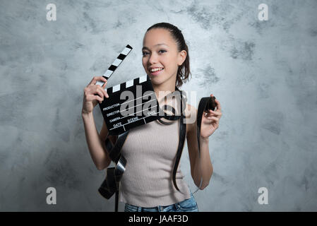 Giovane bella bruna teen donna sorridente ritratto mentre utilizzando movie clapper board e con 35 mm di pellicola intorno al suo corpo su uno sfondo grigio Foto Stock