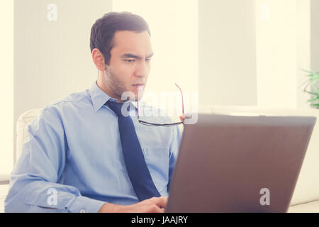 Accigliata uomo morde i occhiali e utilizzando un laptop seduto su un divano Foto Stock