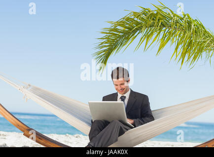 Imprenditore sorridente con notebook e in seduta amaca in spiaggia Foto Stock