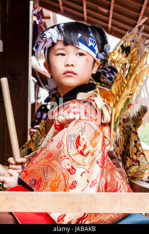 Asian, bambino giapponese, ragazzo, molto viso carino, in seduta Dashi galleggiante tenendo un paio di bastoncini di tamburo. Indossa il kimono arancione e la fascia di testa. Foto Stock