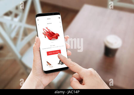 Online store app utilizzare sul moderno telefono cellulare con bordi arrotondati. Donna toccare Aggiungi al carrello pulsante per red shoes. Foto Stock