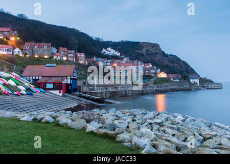 Le luci sono accese nelle case in vista serale del grazioso e pittoresco borgo marinaro, con scogliere, barche & mare calmo - Runswick Bay, North Yorkshire, Inghilterra, Regno Unito. Foto Stock